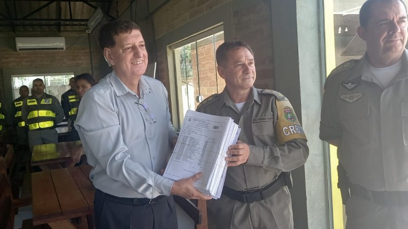 Foto horizontal colorida mostra três homens brancos de cabelos curtos sorrindo. Os dois homens da direita estão com o uniforme da Polícia Militar . O homem à esquerda veste uma camisa social azul e entrega uma pilha de papel para o conselheiro empossado