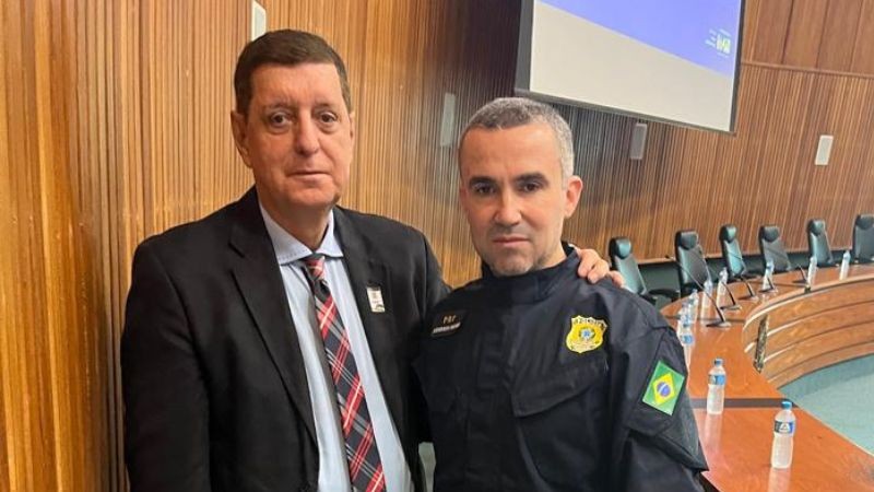 Foto horizontal colorida mostra dois homens brancos olhando para a camera. O da esquerda veste um termo escuro e o da direita, o uniforme azul da PRF.